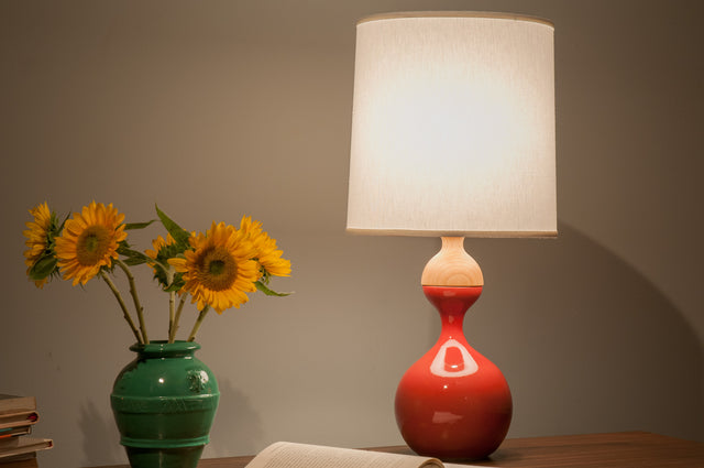 Lit ceramic J Schatz table lamp with a red gourd-shaped base and a white linen shade, beside sunflowers in a green vase