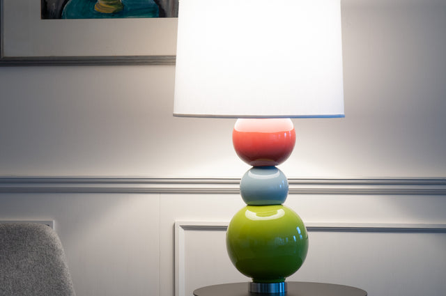 Lit ceramic J Schatz table lamp with stacked coral, pale blue and chartreuse glazed spheres under a white shade, against a panelled wall