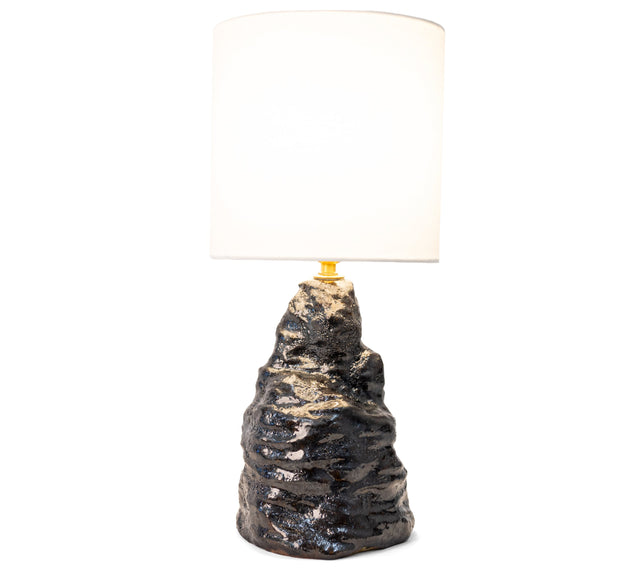 Amorphous table lamp 1, a broad craggy stoneware base in black metallic lava glaze under a fabric shade, by Jim Schatz, J Schatz