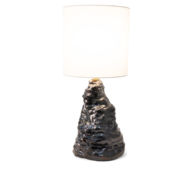 Amorphous table lamp 2, a tapering craggy stoneware base in black metallic lava glaze under a fabric shade, by Jim Schatz, J Schatz